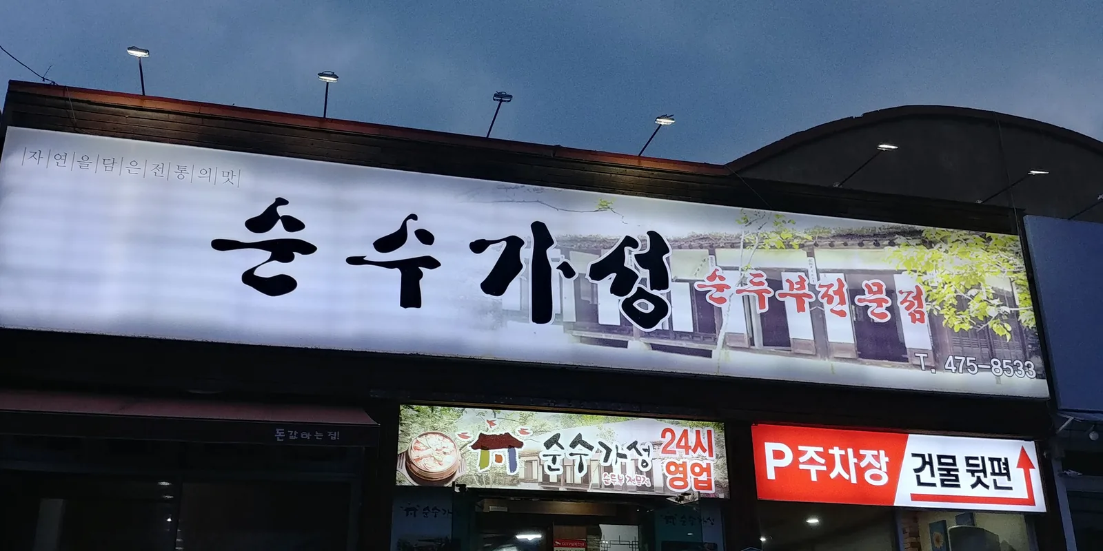 저녁의 가게 간판 — 불 켜진 큰 간판 «순수가성 순두부전문점», 입구 위 «24시 영업» 판, 오른쪽 «P 주차장 건물 뒷편» 안내